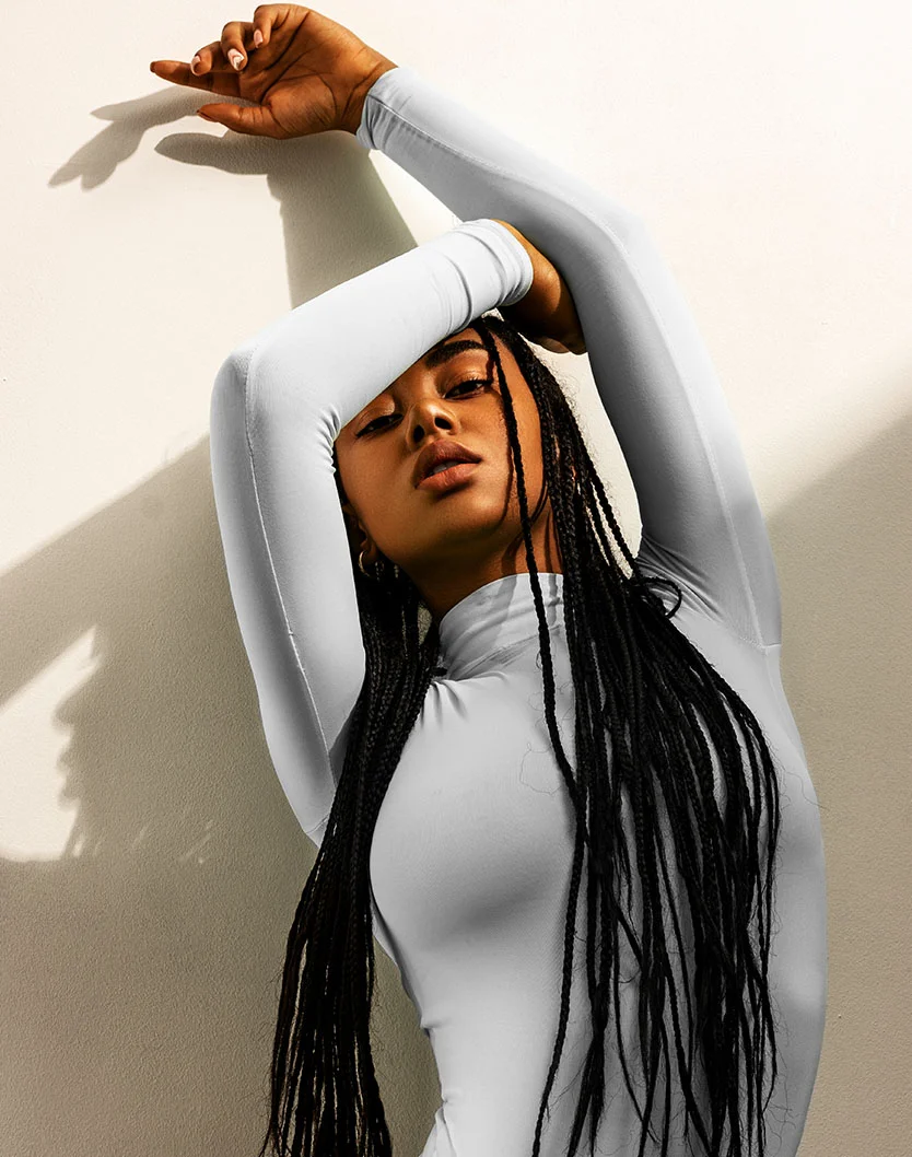 A young woman with dark braided hair poses against a light beige wall. She wears a form-fitting, long-sleeved turtleneck in light gray or off-white. One hand lightly touches her forehead while the other rests at her side. Her neutral expression and direct gaze add to the artistic, fashion-inspired feel. Soft lighting casts subtle shadows, accentuating her form and the contrast between her dark hair and the light background. - Breast Implant Exchange Capsular Contraction Recovery in Las Vegas, NV