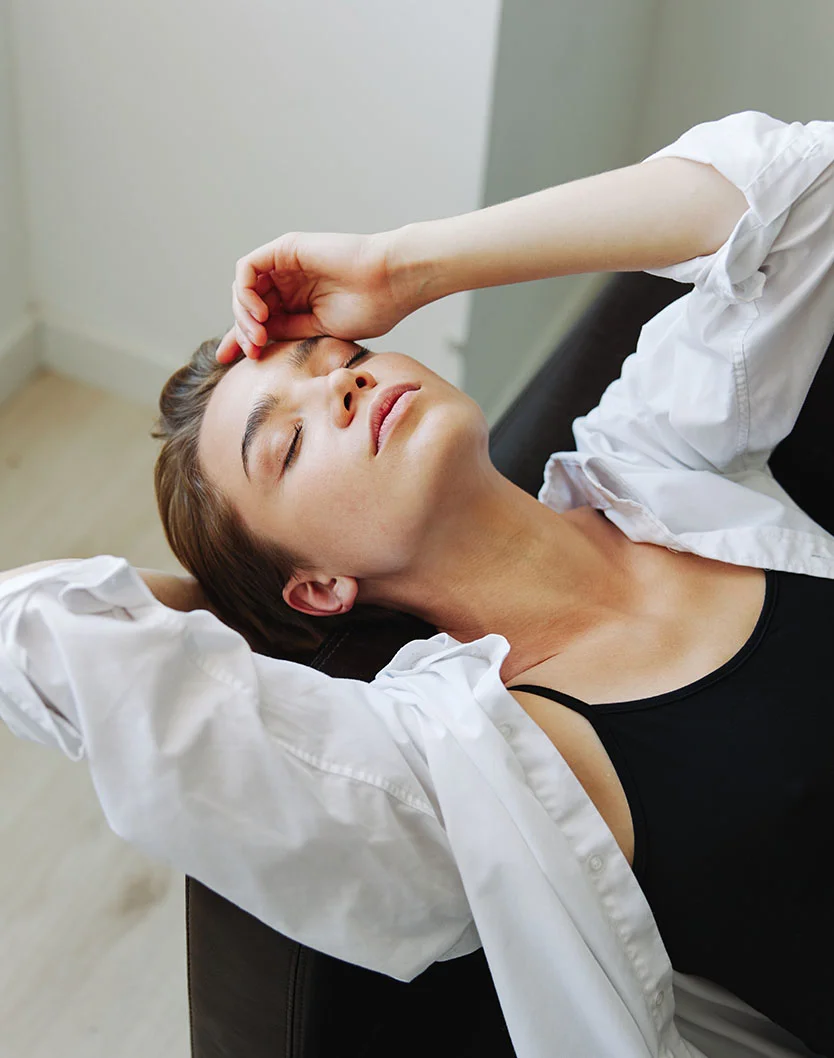 A young woman is reclining with her head resting in her hand, eyes closed, and a relaxed expression. She wears a loose white long-sleeved shirt over a black tank top. Her dark brown, slightly wavy hair falls around her shoulders. The background is a neutral-toned room with dark grey/black furniture beneath her. Soft, diffused lighting adds to the calm, relaxed atmosphere. - Browlift Recovery in Las Vegas, NV