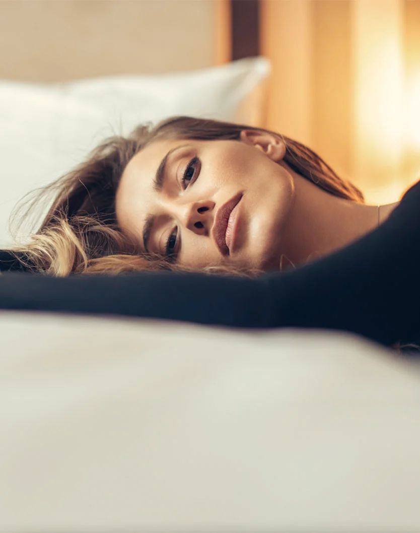 Close-up of a woman lying on a bed with shoulder-length light brown hair. She has a relaxed expression, her eyes looking downward, and her lips closed. Her smooth, fair skin is highlighted by soft lighting. She wears a dark top, and the white bed with smooth, crisp sheets creates a serene atmosphere. The background suggests a bedroom setting with diffused lighting. - Eyelid Surgery Recovery in Las Vegas, NV