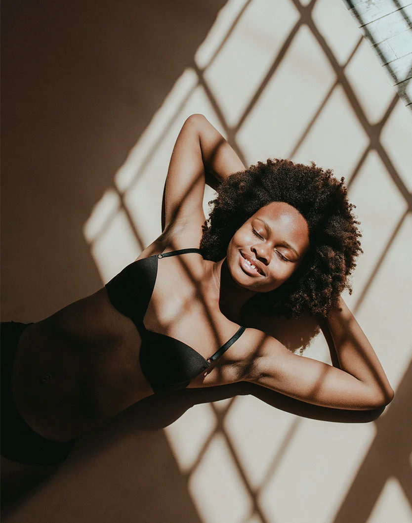 A woman with dark brown curly hair is lying on a light beige floor or wall. She's wearing a black bra and underwear. Her arms are outstretched, and her head is turned slightly to the right. Her eyes are closed, and she appears to be relaxed. The light from a window with a diamond-patterned grid creates strong shadows on the floor or wall beneath her and around her body. The overall style of the image is soft and natural. - Body Lift at LV Body Sculpting