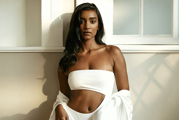 A woman with brown skin and long, dark brown hair is pictured from the mid-chest up, standing against a light beige wall with a white window frame above her. She is wearing a white, ribbed, strapless top, and a white, draped garment is loosely positioned around her lower body. Her expression is neutral as she looks straight ahead. Soft, natural light casts gentle shadows behind her, creating a calm and minimal portrait atmosphere. - Breast Augmentation at LV Body Sculpting