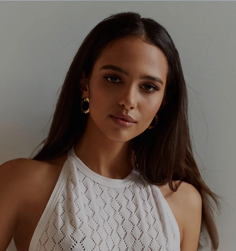 A woman with long, dark brown hair and light brown skin looks directly at the camera with a neutral expression. She is wearing light makeup and natural pink lipstick. Gold-toned, textured hoop earrings accent her look. Her top is a white, lace-like or knit halter style with a diamond-patterned texture. The background is a muted, light gray, and the soft, diffused lighting highlights her features evenly. - Contour and Define at LV Body Sculpting