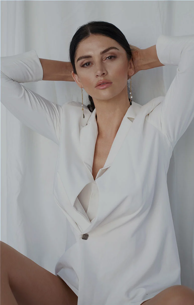A woman with light tan skin and dark brown or black hair is seated, wearing a white, long-sleeved blazer-style top with a draped, sculpted V-neck opening. Her arms are raised, hands resting lightly on the back of her head. She has long earrings with small, light-colored stones or beads. Her hair is pulled back in a smooth, slightly loose style. The background is a soft cream or beige tone, and the lighting is even and neutral. Her posture is relaxed yet composed, giving an impression of calm confidence. - Lift and Rejuvenate at LV Body Sculpting