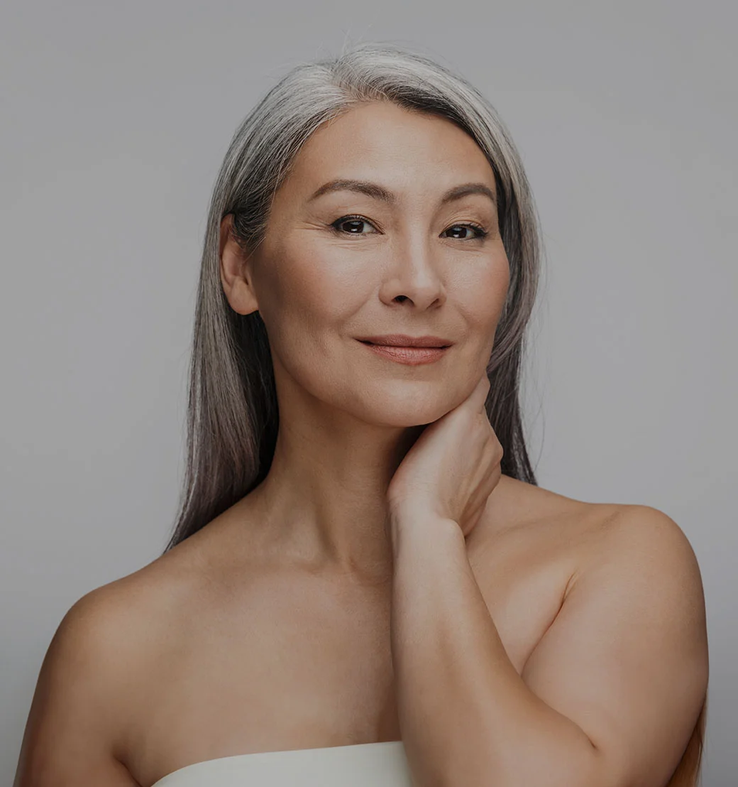 A medium shot of a middle-aged woman with a serene expression. She has shoulder-length, graying hair and a light beige/tan complexion. Her skin appears smooth and soft. Her hand rests gently on her neck. She is wearing a simple, off-white strapless top. The background is a neutral gray. The focus is on her face and upper body, conveying a sense of calmness and well-being. - Restore Hair at LV Body Sculpting